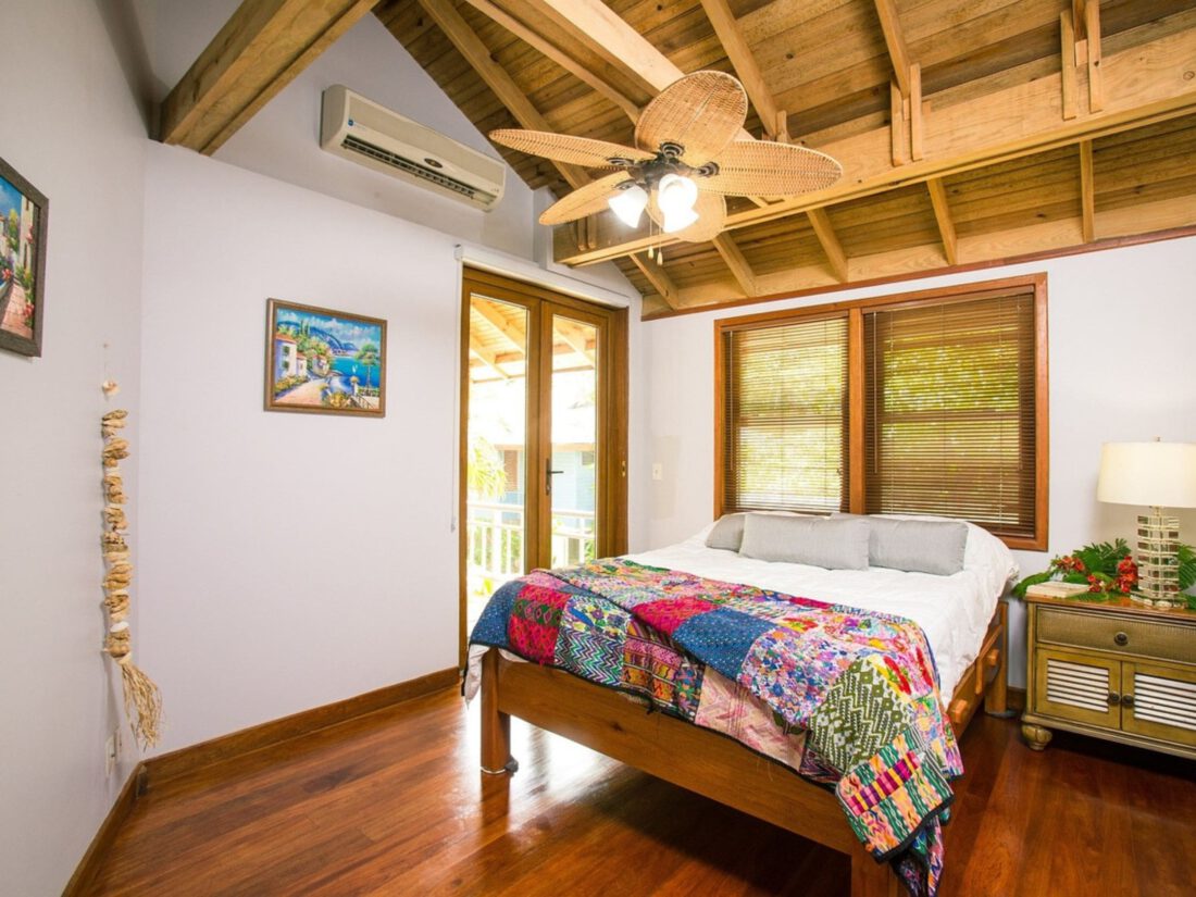 "Immagine di una camera di una casa vacanze arredata con stile marino. Letto matrimoniale con biancheria dai toni del blu e bianco, abbinata a complementi d'arredo in rattan e bambù. La stanza è luminosa e ben organizzata, con mobili multifunzionali che ottimizzano lo spazio. Una finestra aperta lascia entrare luce naturale e una brezza fresca, mentre decorazioni ispirate al mare, come conchiglie e quadri di paesaggi costieri, creano un'atmosfera rilassante e accogliente. Un angolo lettura con poltrona in tessuto lavabile e lampada da terra invita al relax dopo una giornata al mare