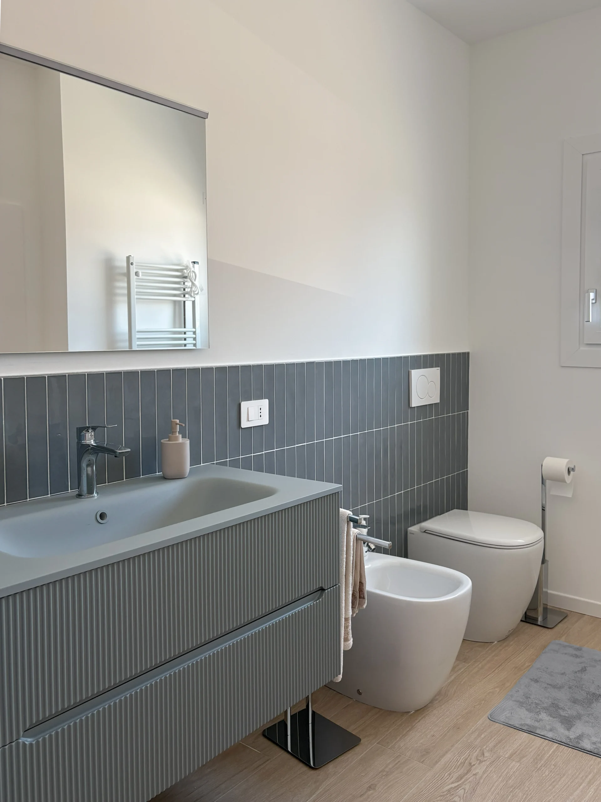 Bagno moderno con mobile lavabo sospeso grigio, frontali scanalati, rivestimento coordinato, sanitari bianchi e ampio specchio.