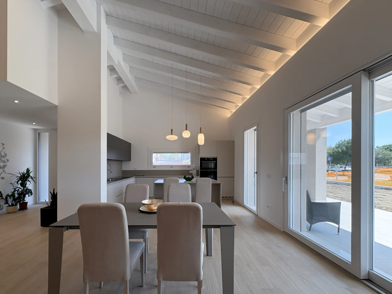 Zona giorno luminosa con cucina moderna su misura, sala da pranzo, soffitto con travi bianche e ampie vetrate affacciate sul portico.
